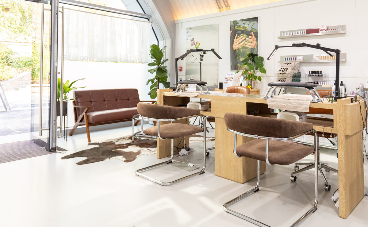 A modern nail salon interior with multiple wooden manicure stations, brown upholstered rolling chairs, shelving, and large windows with an outdoor patio visible.